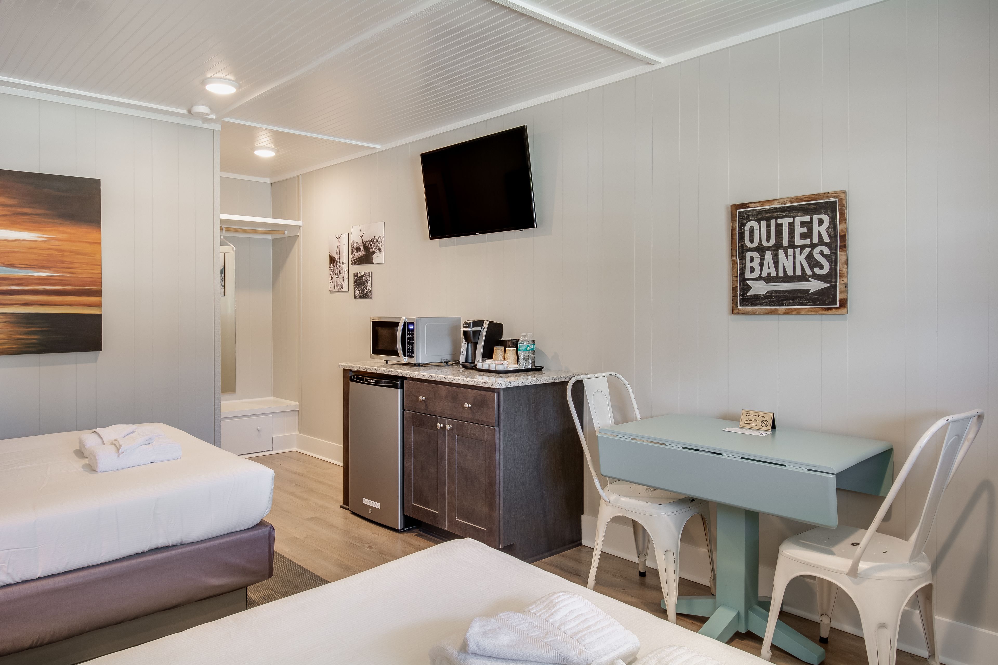 A hotel room with two beds, a desk, a TV, and a sign indicating 'Outer Banks'.