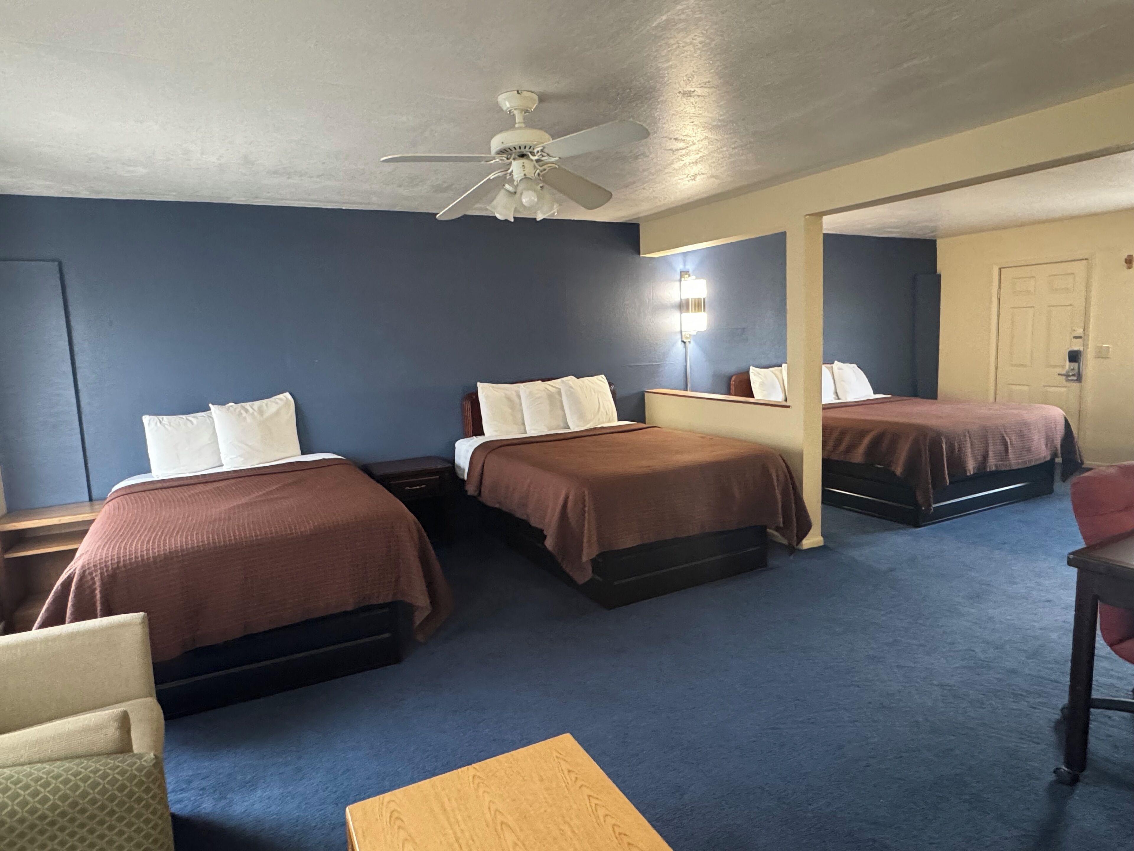 Una habitación de hotel con tres camas, un ventilador de techo, una mesita de noche, una silla y una pequeña mesa.