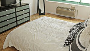 A bedroom with a large bed, a dresser, a television, a mirror, and a window with blinds. - BellaView Suites (Jersey City)
