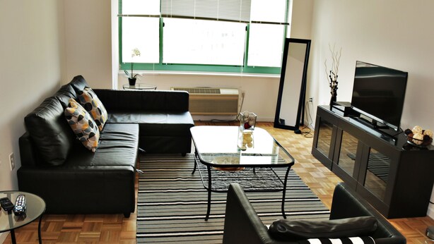 A modern living room with a black leather sofa, a glass coffee table, a black armchair, and a television on a cabinet. - BellaView Suites (Jersey City)