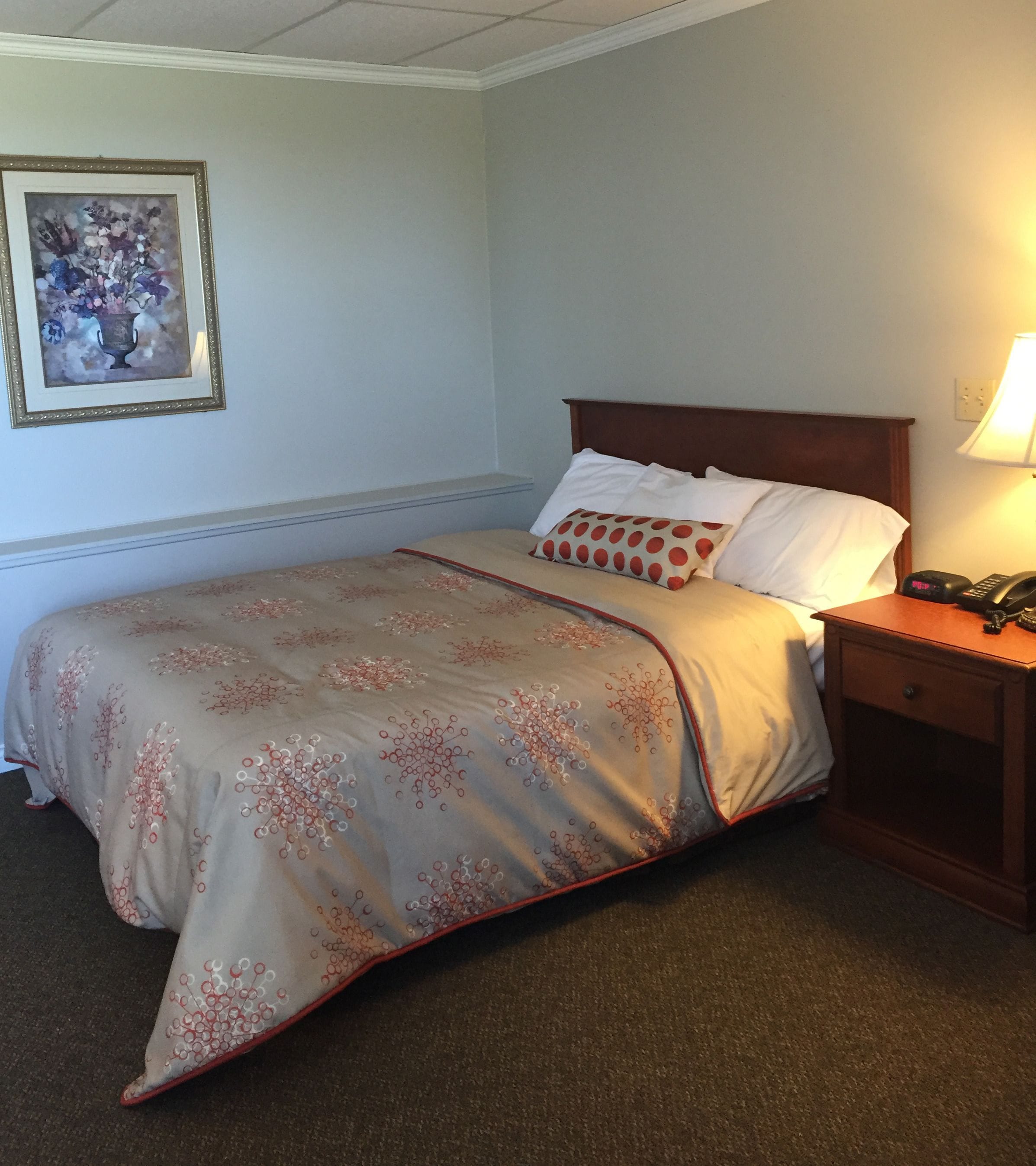 A neatly made bed with a floral-patterned bedspread, a wooden headboard, and a bedside table with a lamp and a phone.