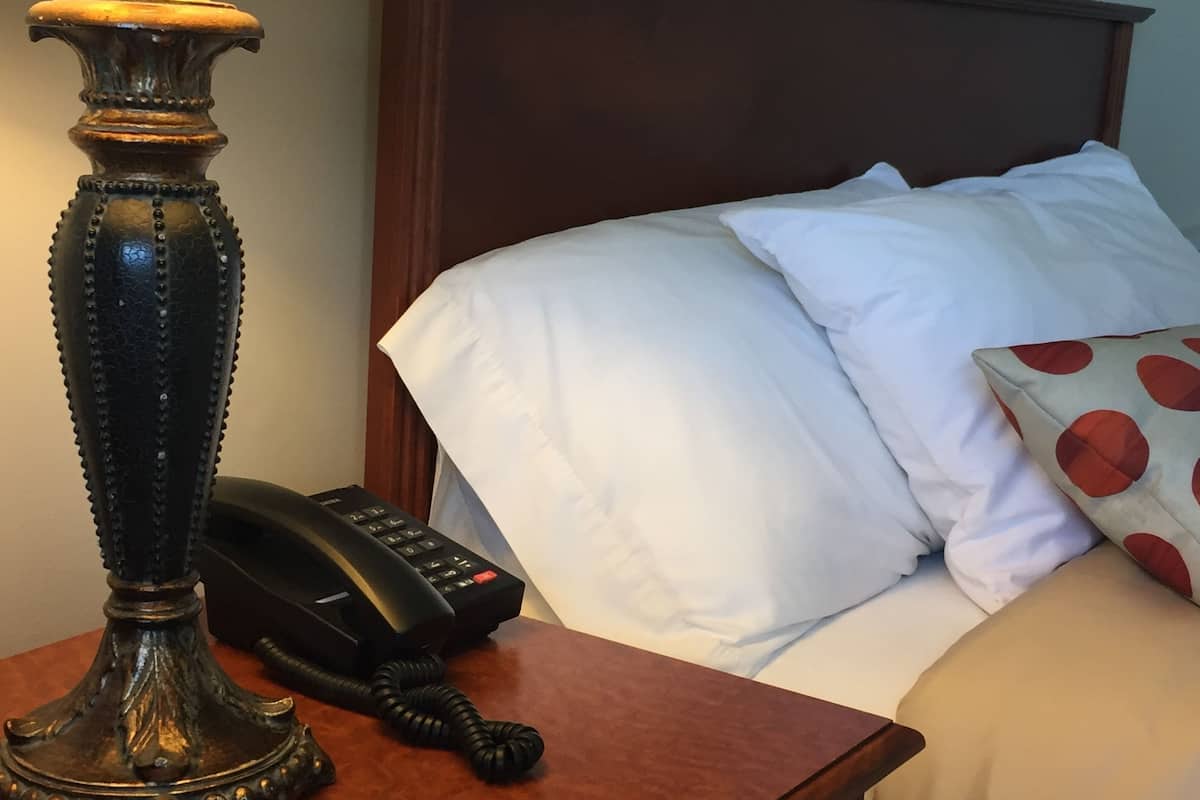 Una mesita de noche de madera con una lámpara, un teléfono y una cama con almohadas blancas y estampadas.