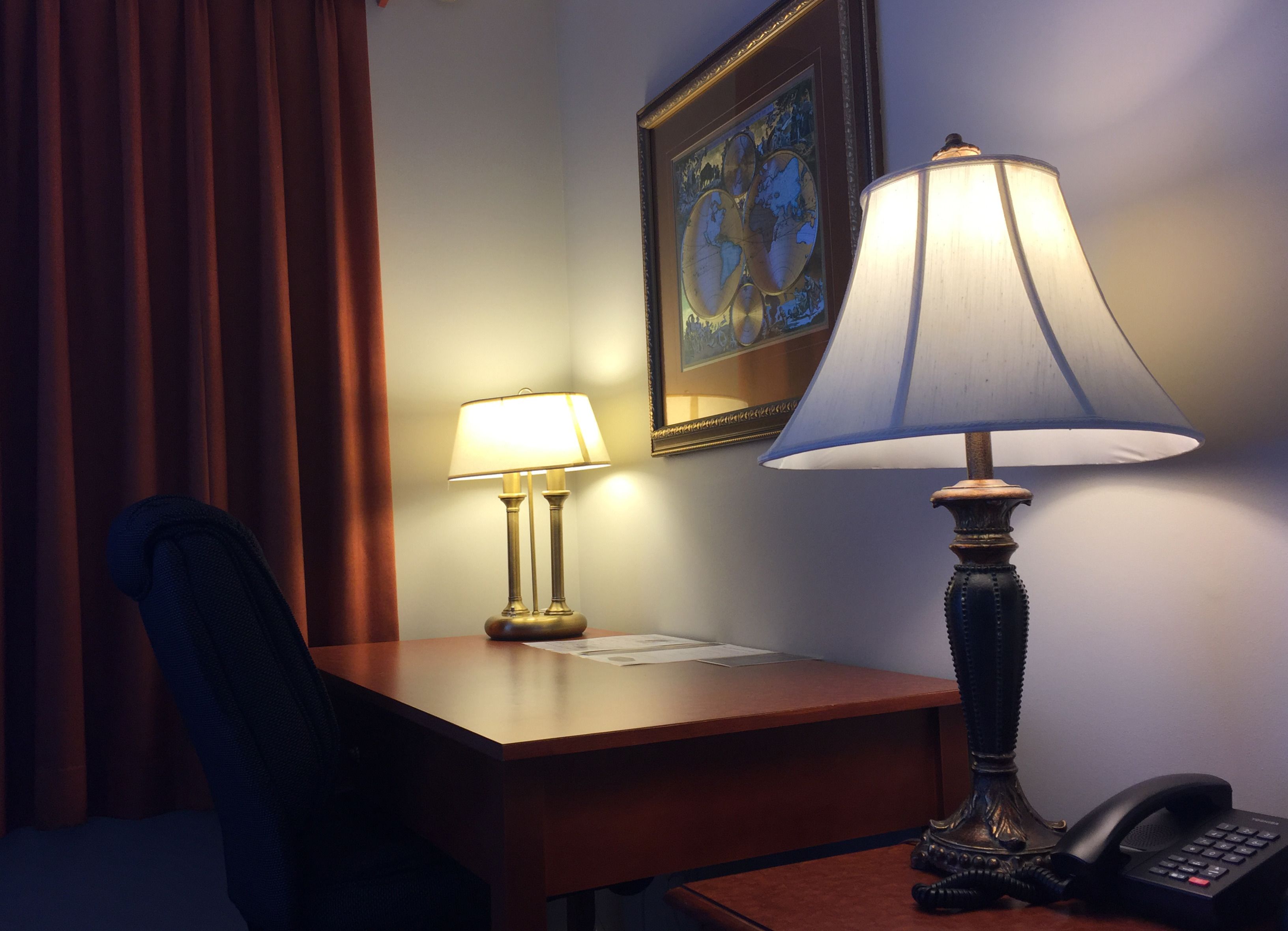 Et hotelværelse med et skrivebord, to lamper, en stol, et indrammet verdenskort og en telefon.