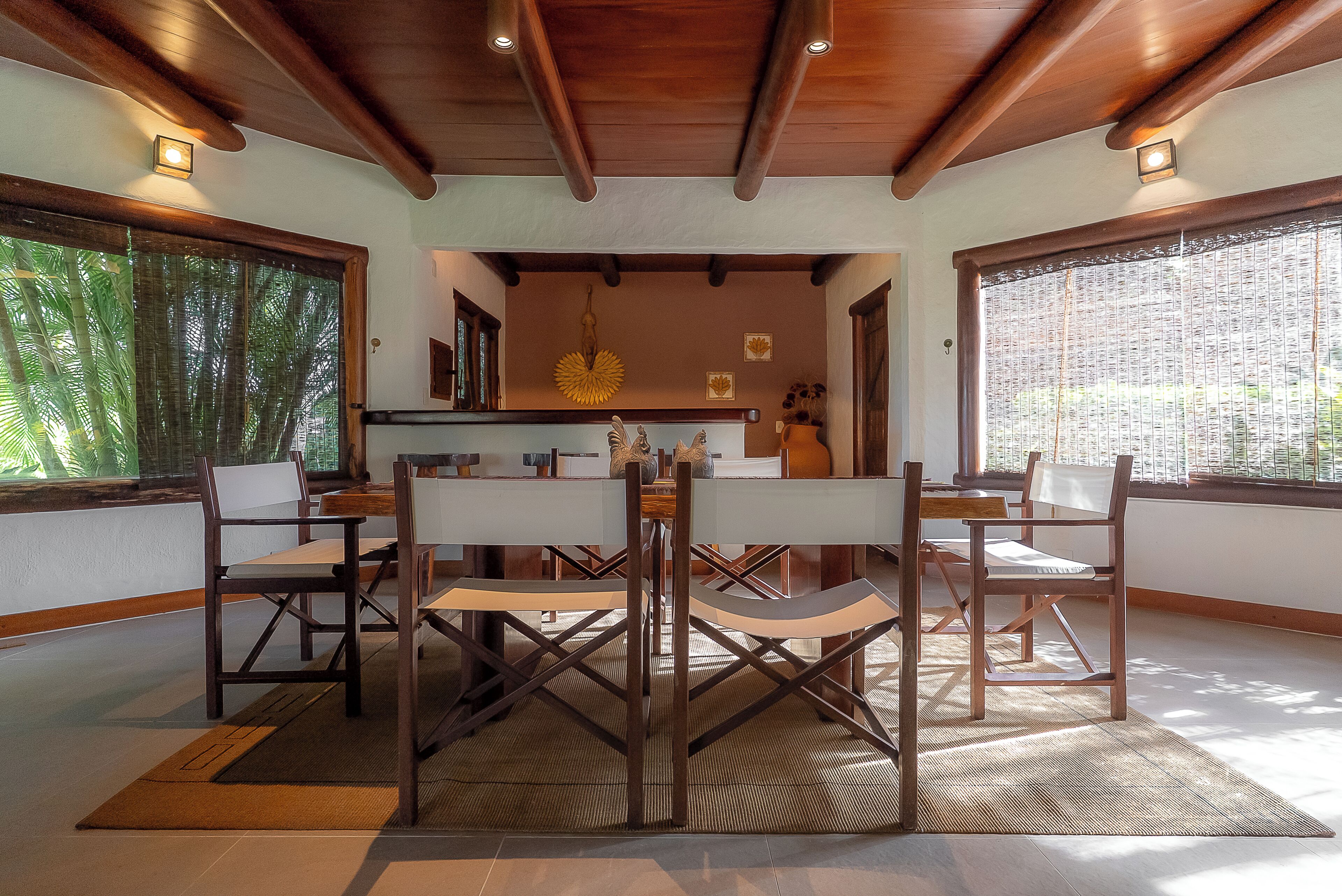 A dining area with a wooden table and chairs, a central light fixture, and large windows with a view of greenery.