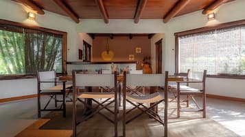 A dining area with a wooden table and chairs, a central light fixture, and large windows with a view of greenery.