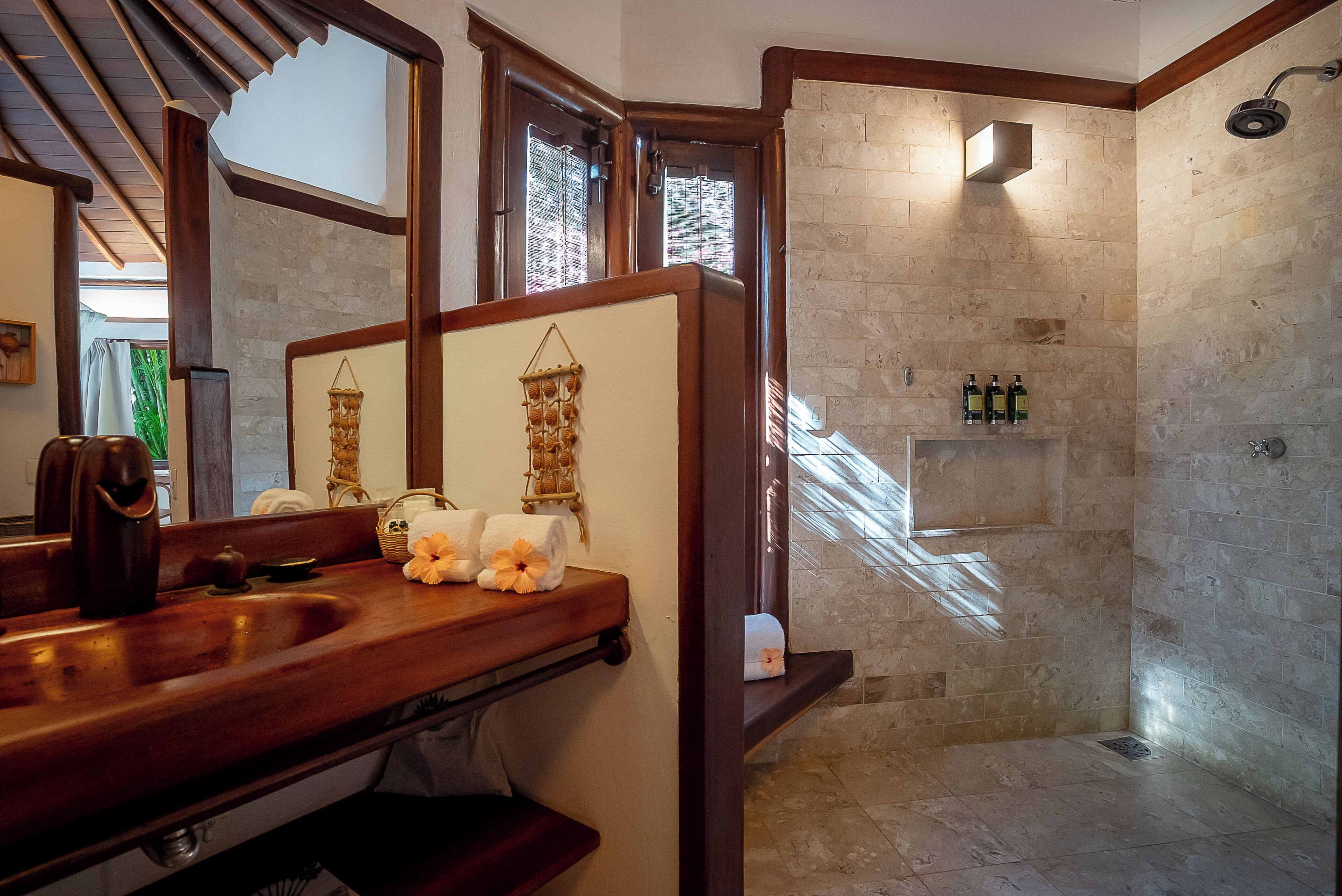 Casa de banho com toucador de madeira, um espelho grande, chuveiro com azulejos de pedra e uma prateleira com produtos de higiene pessoal.