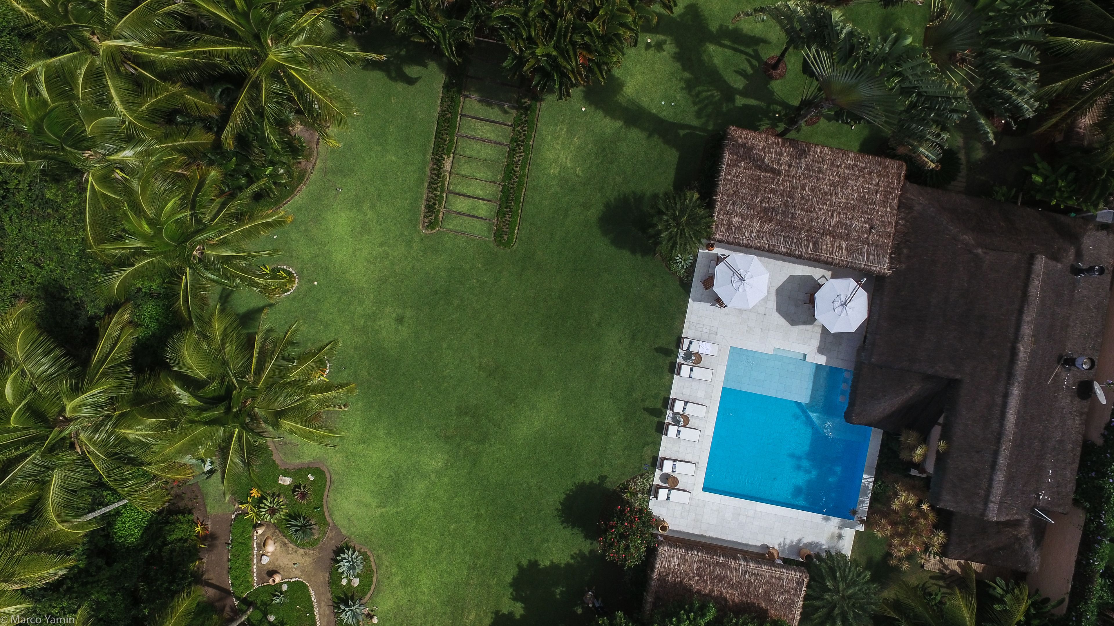Vista aérea de una propiedad con piscina, zona de patio con sombrillas y césped bien cuidado rodeado de palmeras.