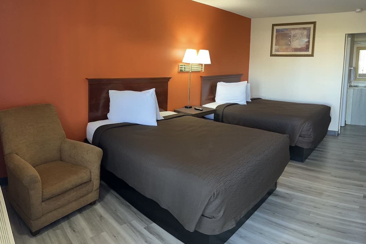 A hotel room with two beds, a chair, a lamp, and a painting on the wall.