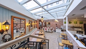 Un restaurant avec un plafond haut, des murs en briques apparentes et un toit en verre. Les tables et les chaises sont disposées pour accueillir les clients.