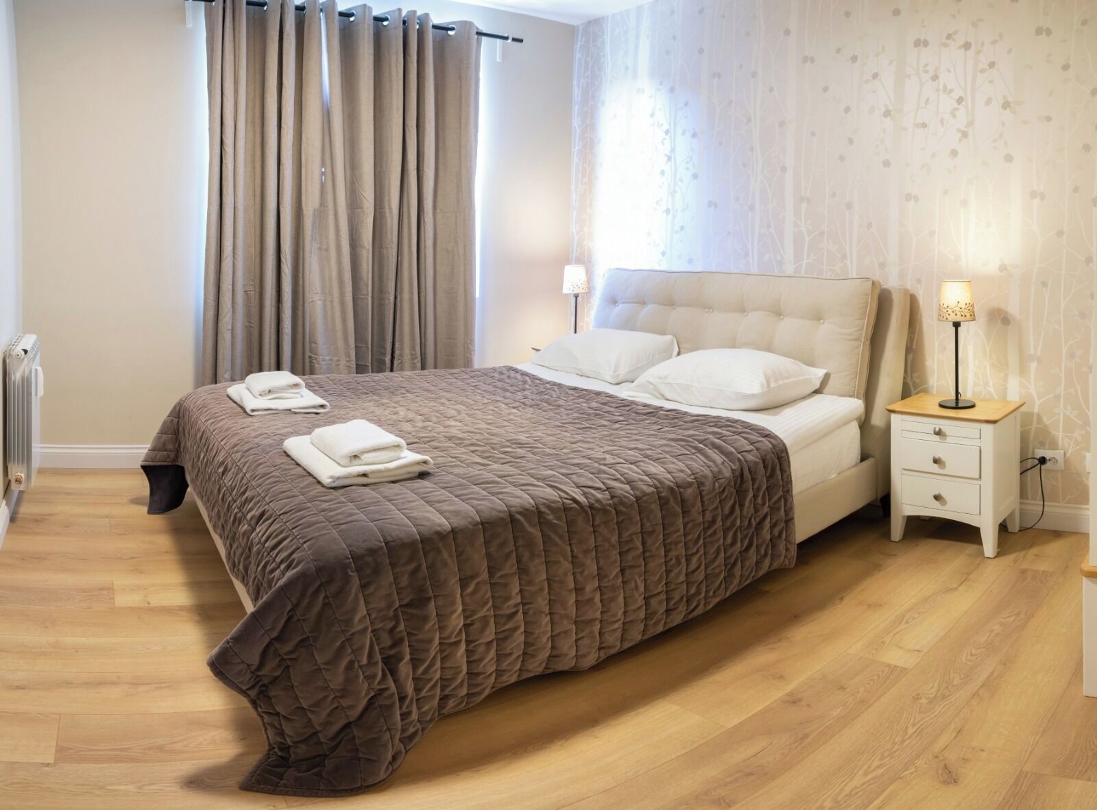 A neatly made bed with a brown quilt, white pillows, and folded towels on the side. There is a bedside table with a lamp and a small drawer. The room has wooden flooring and a radiator on the wall.