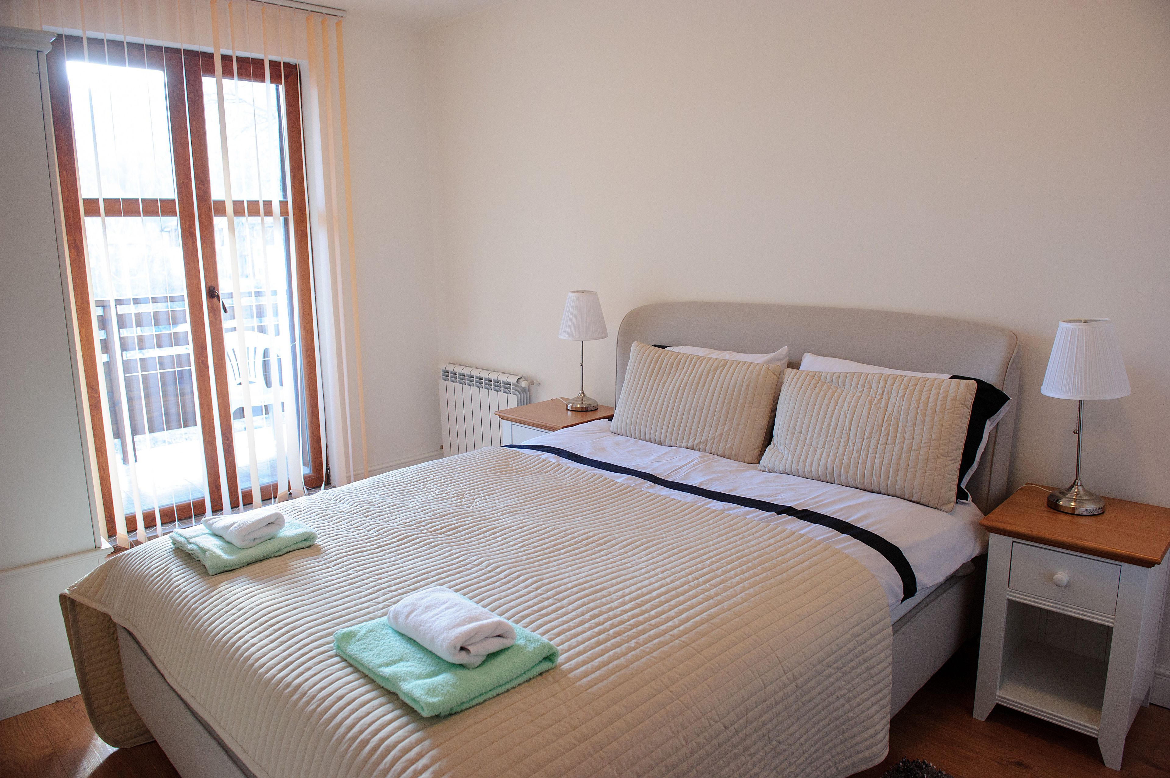 A neatly made bed with a quilted cover, two bedside lamps, and a window with blinds.