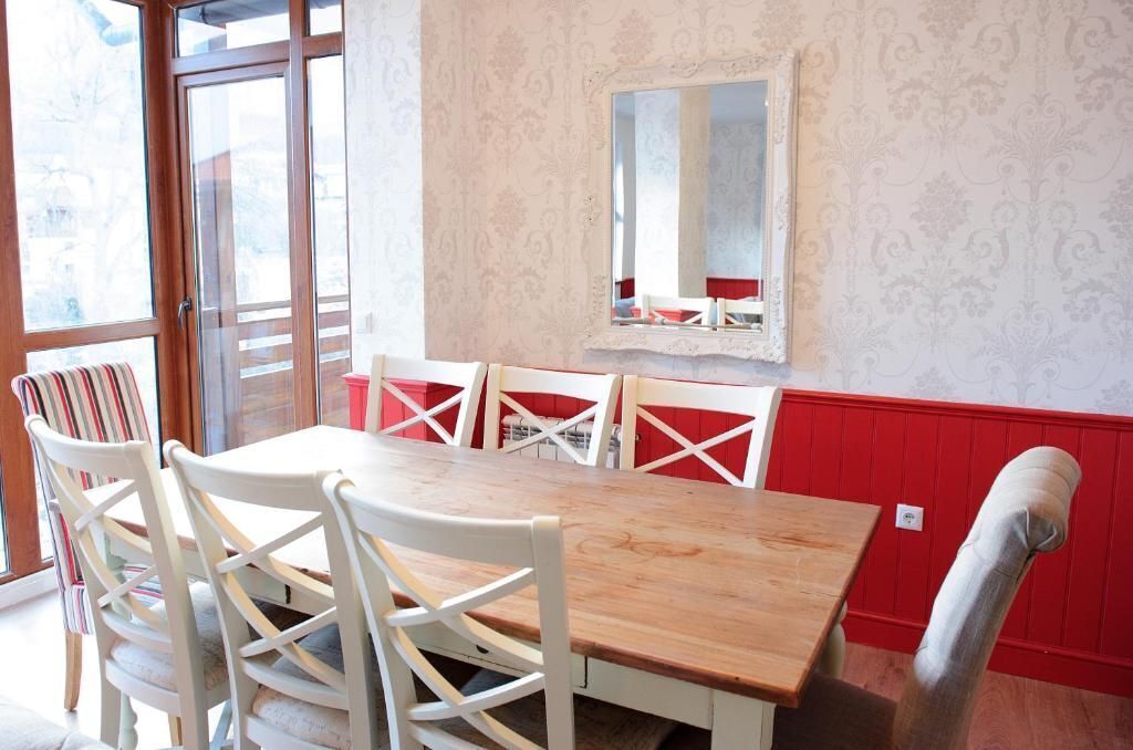 Un comedor con mesa de madera y sillas blancas, un banco tapizado en rojo y un espejo en la pared.