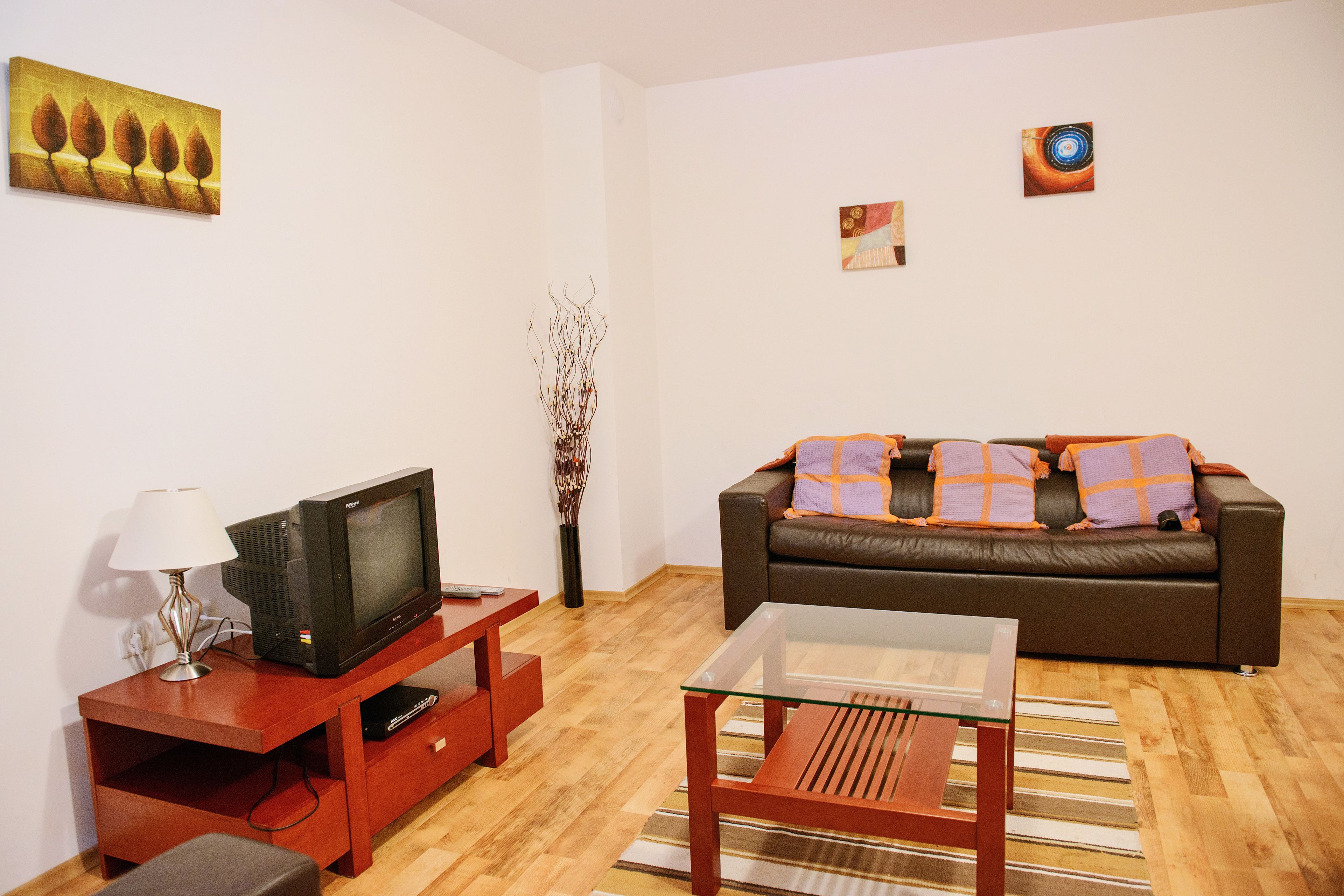Una sala de estar con un sofá de cuero marrón, una mesa de centro de vidrio, un mueble de madera para televisión y una lámpara.