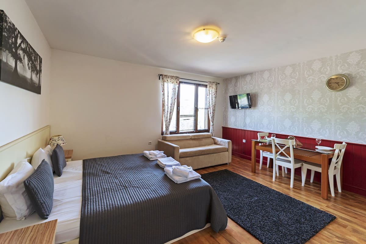 A hotel room with a bed, a sofa, a dining table with chairs, a TV mounted on the wall, and a window with curtains.