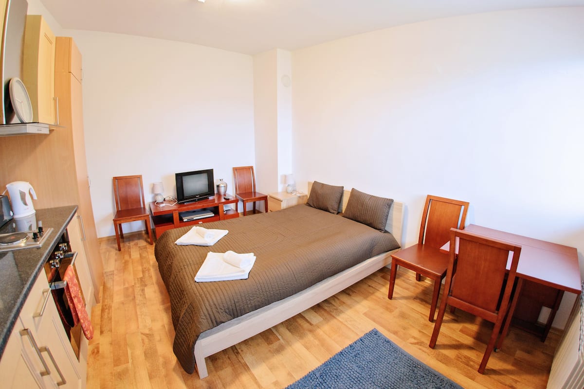 A bedroom with a bed, a desk, a chair, a television, and a kitchenette.
