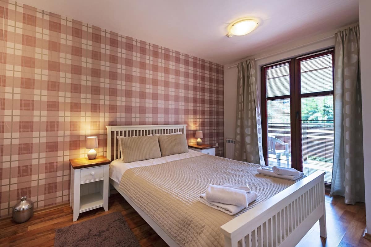 A bedroom with a checkered wall, a bed with a headboard, bedside lamps, a window with blinds, and a radiator.