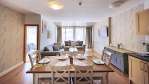A dining area with a wooden table set for four, surrounded by white chairs. There is a sofa and a television in the background. - Mountain View Resort (Bansko)