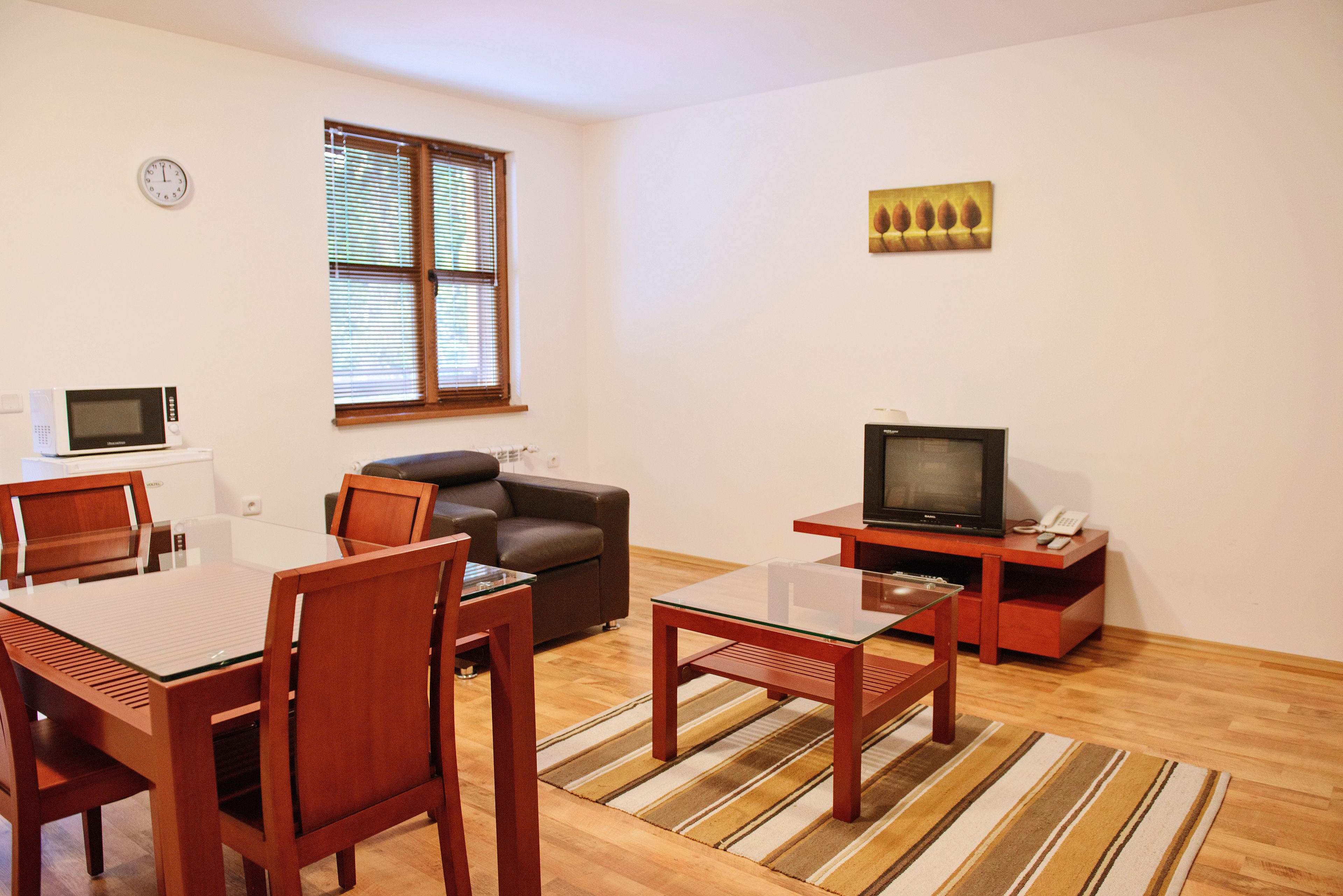Una sala de estar con una mesa de comedor, sillas, un sofá, un televisor, un reloj y una ventana con persianas.