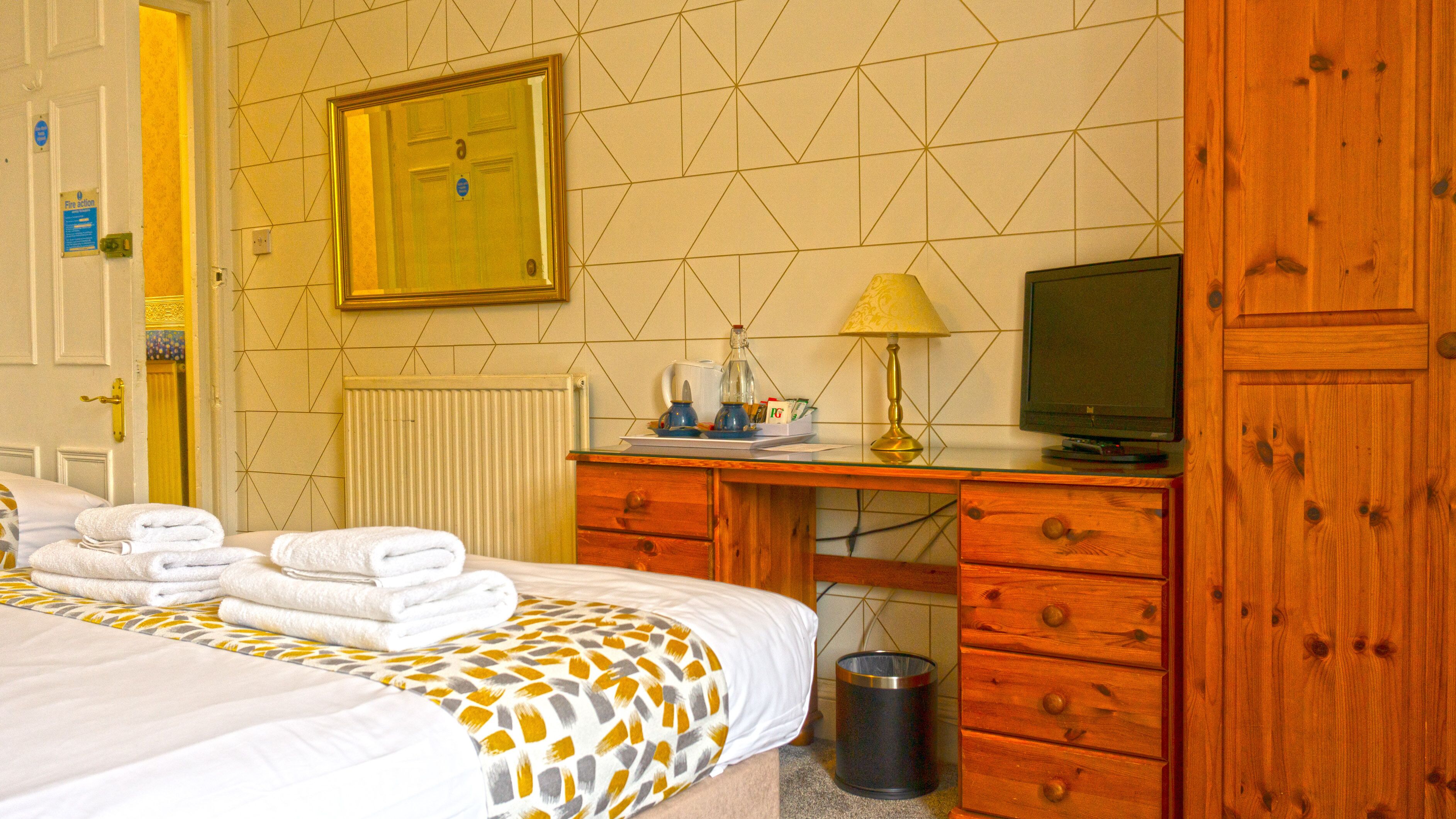 Habitación de hotel con cama, escritorio con lámpara, televisión, radiador y cubo de basura.