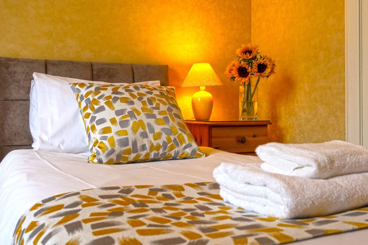 A neatly made bed with a patterned bedspread, a pillow, and two folded towels. There is a bedside lamp and a vase with sunflowers on a nightstand.