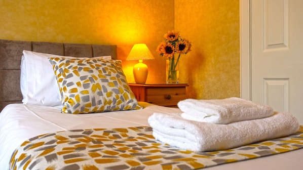 A neatly made bed with a patterned bedspread, a pillow, and two folded towels. There is a bedside lamp and a vase with sunflowers on a nightstand. - Arrandale Guest House (Edinburgh)