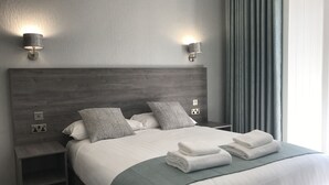 Ein modernes Hotelzimmer mit einem Holz-Kopfteil, einem ordentlich bezogenen Bett mit weißen Laken und grauen Kissen, zwei Nachttischlampen und Blick ins Freie durch ein mit Vorhängen versehenes Fenster.