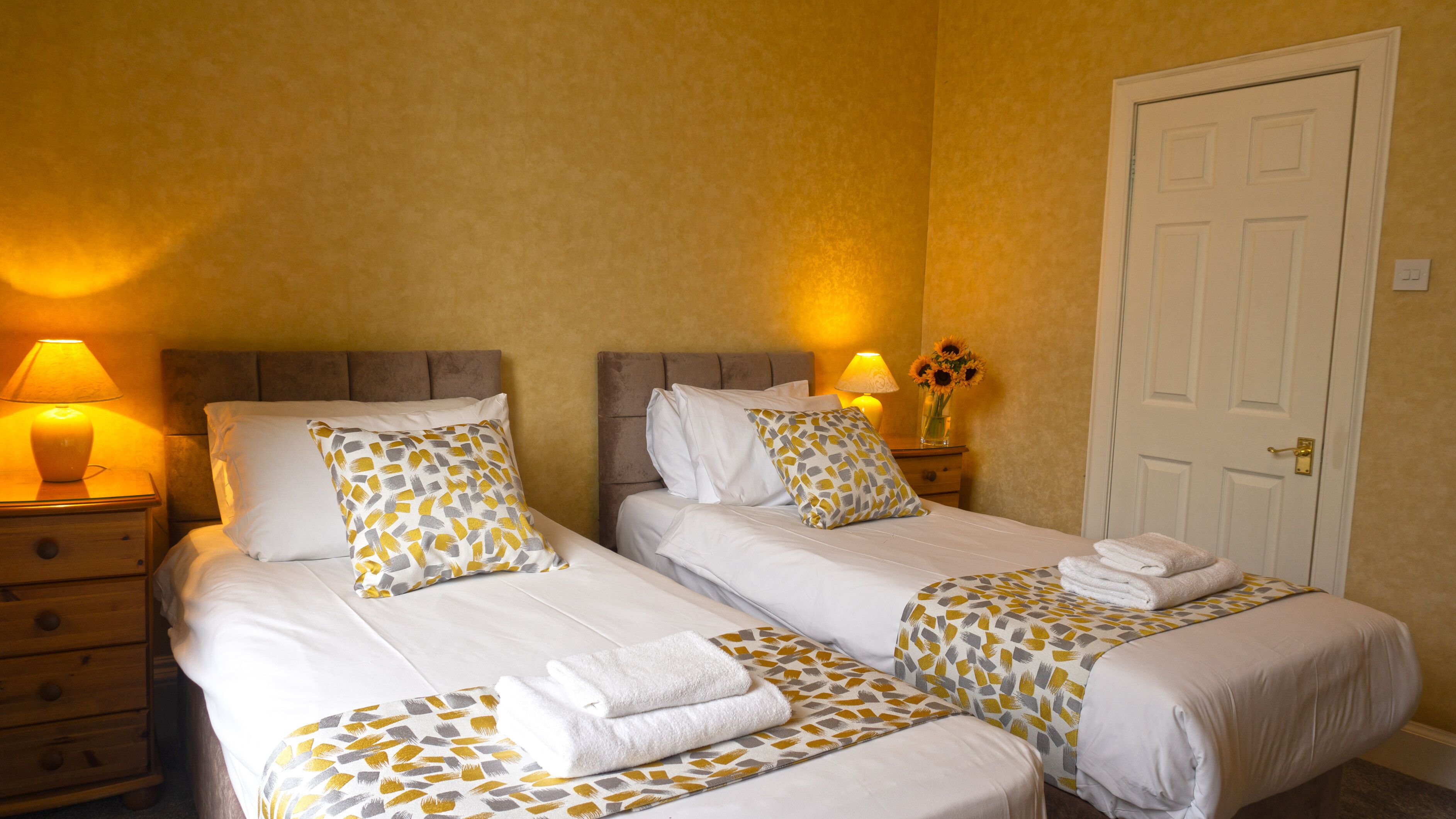 Een hotelkamer met twee bedden, elk voorzien van een beddekleed met dessin en kussens, een nachtkastje met een lamp, een vaas met bloemen en een deur.