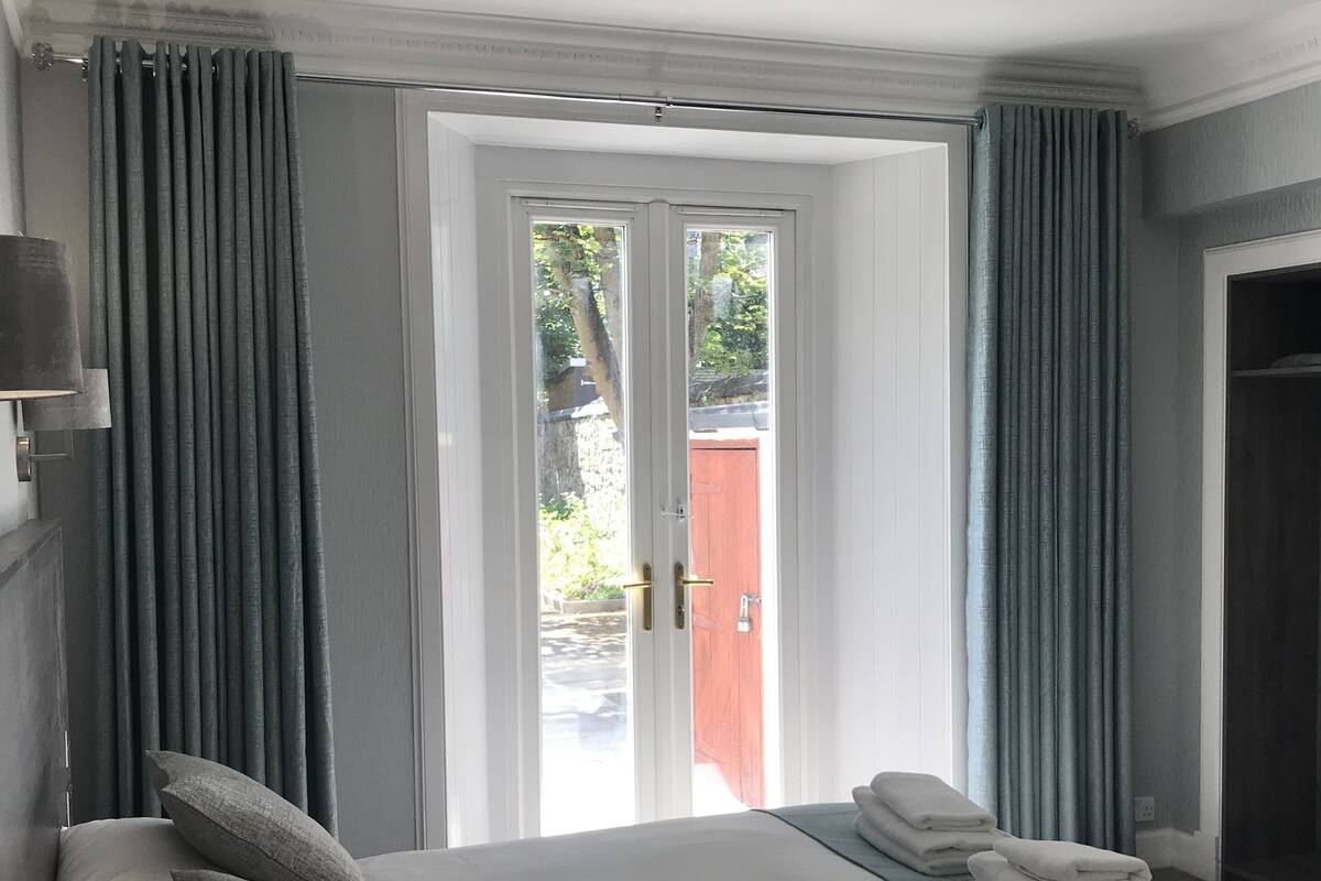 A bedroom with a bed, pillows, a bedside lamp, a window with curtains, and a view of the outside.