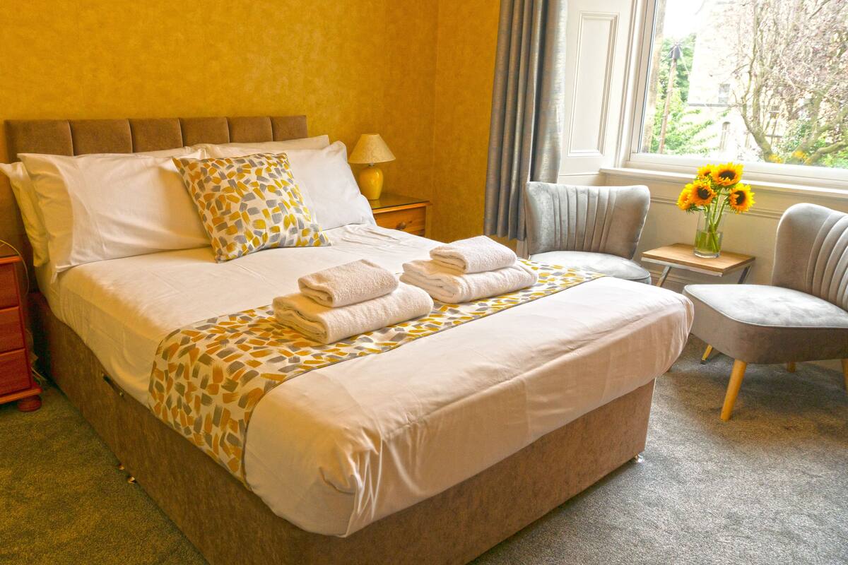 A neatly made bed with towels, a bedside table with a lamp, a chair, and a window with a view of greenery.