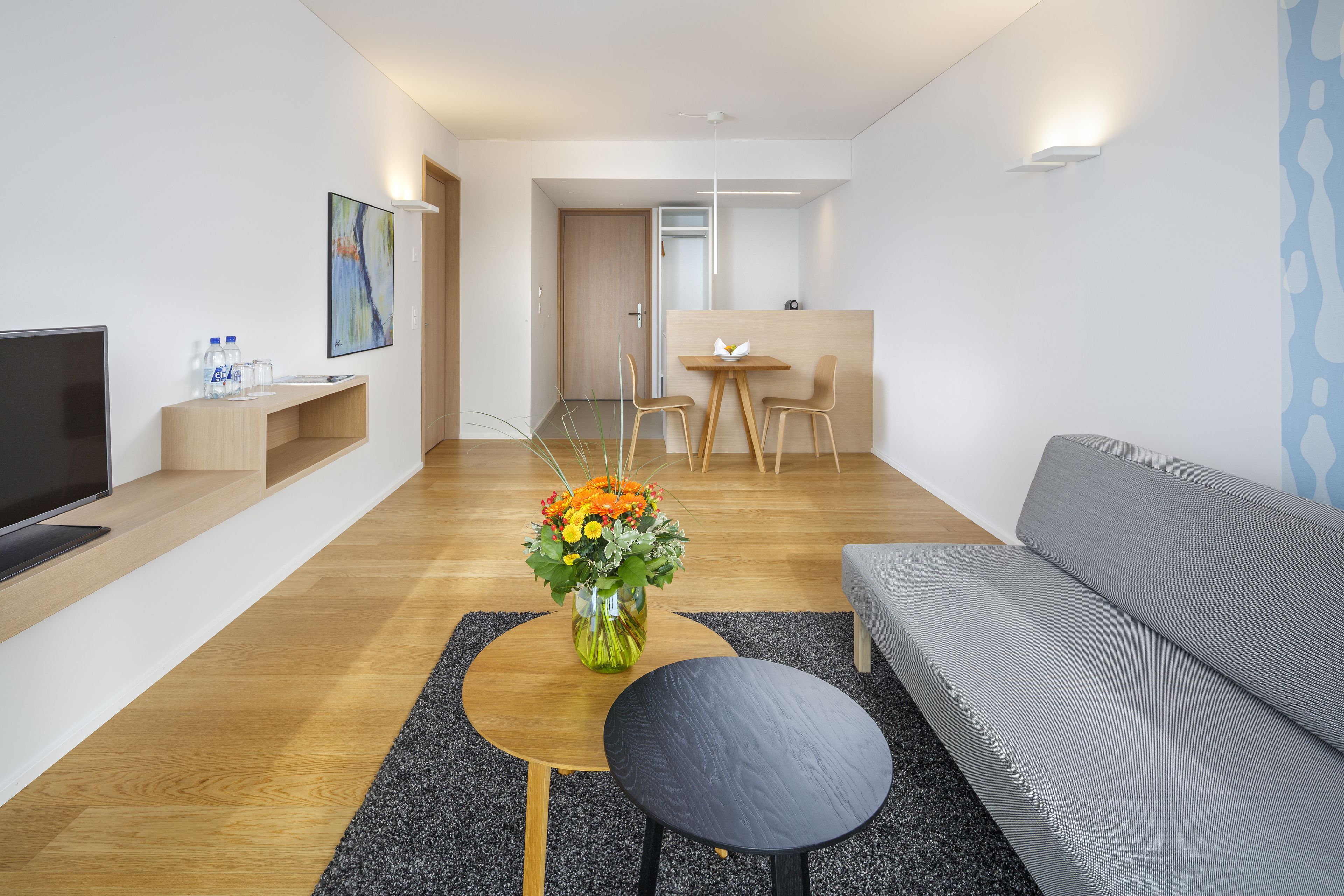 A modern living room with a grey sofa, a wooden coffee table, a black side table, a flat-screen TV, and a vase of flowers.