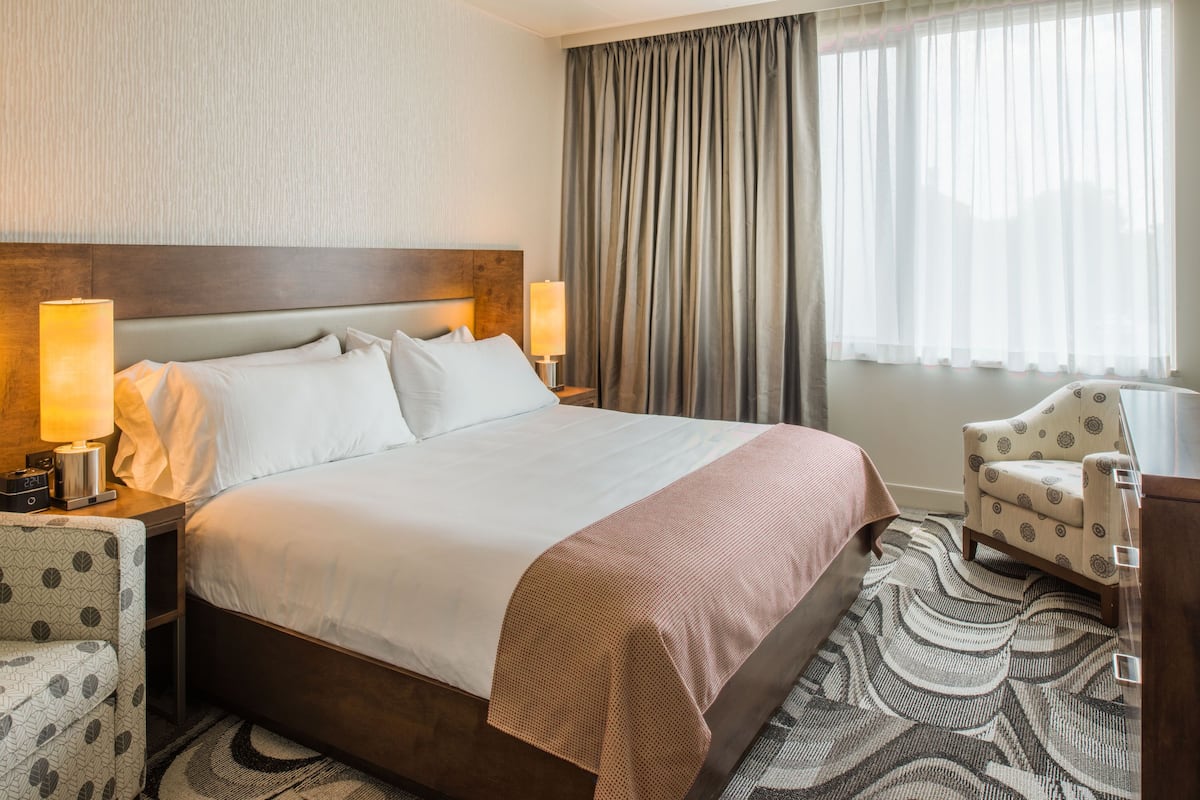 A hotel room with a large bed, two bedside lamps, a patterned armchair, and a view of the outdoors through sheer curtains.