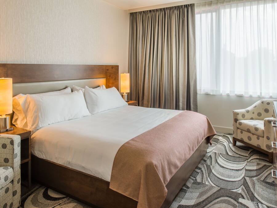 A hotel room with a large bed, two bedside lamps, a patterned armchair, and a view of the outdoors through sheer curtains.