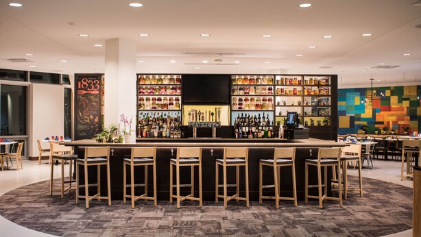 A modern bar area with a central counter, multiple stools, and shelves stocked with bottles. - The Hotel At Oberlin (Oberlin)