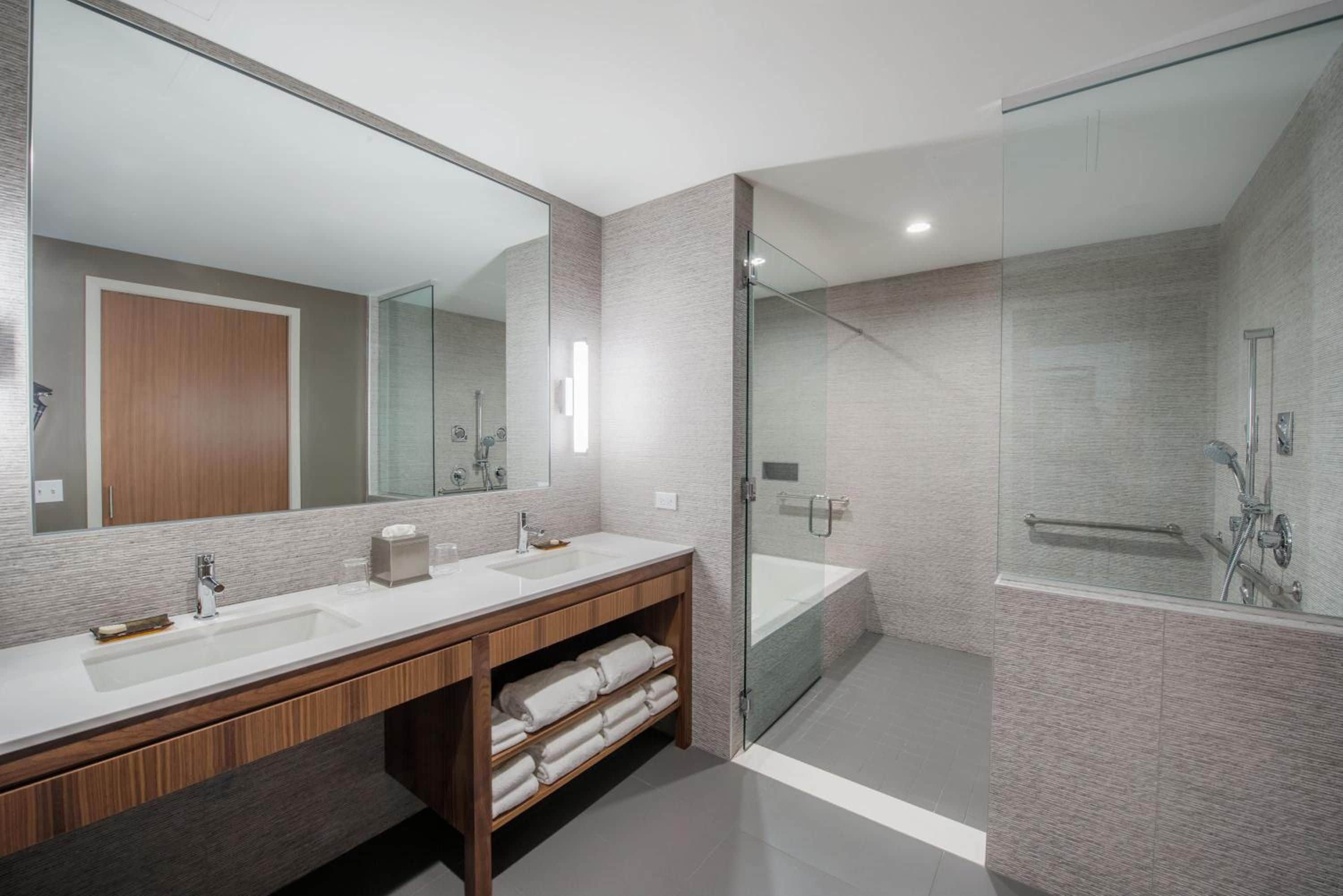 A modern bathroom with a double sink vanity, a large mirror, a glass-enclosed shower, and a wooden cabinet with towels.