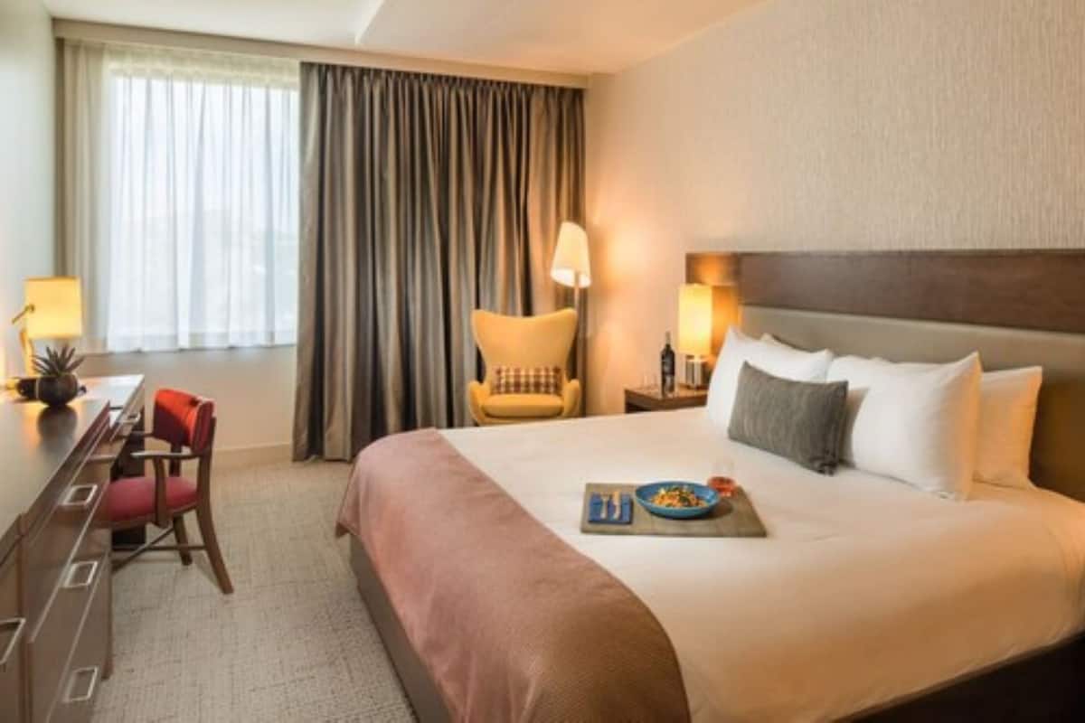 A hotel room with a large bed, a desk with a chair, a lamp, and a view of the outside through a window with curtains.
