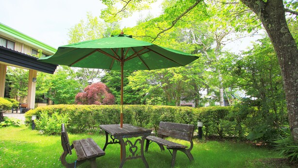 A garden with a wooden table and bench under a green umbrella, surrounded by lush greenery and a building in the background. - Kounso (Yamanakako)