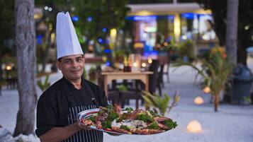 A chef holding a large platter of seafood in an outdoor dining area with tables and chairs, surrounded by trees and ambient lighting.
