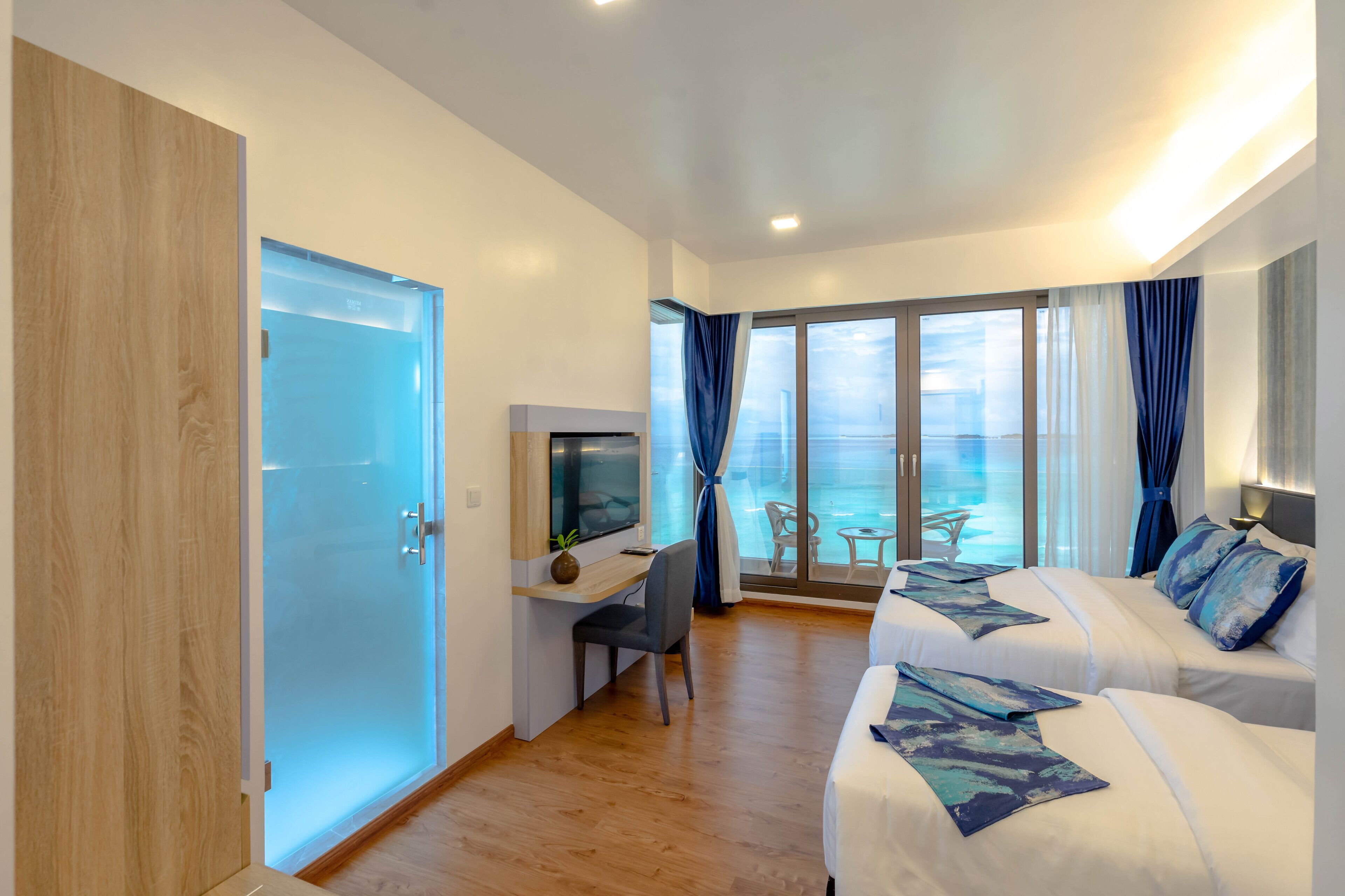 A modern hotel room with a balcony, a desk with a TV, two beds, and a wooden door.