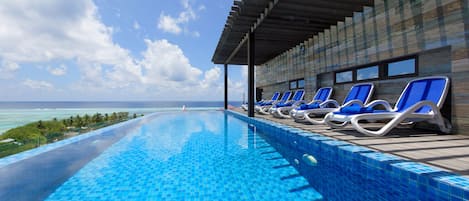 A pool with blue tiles, a covered area with lounge chairs, and a view of the ocean.