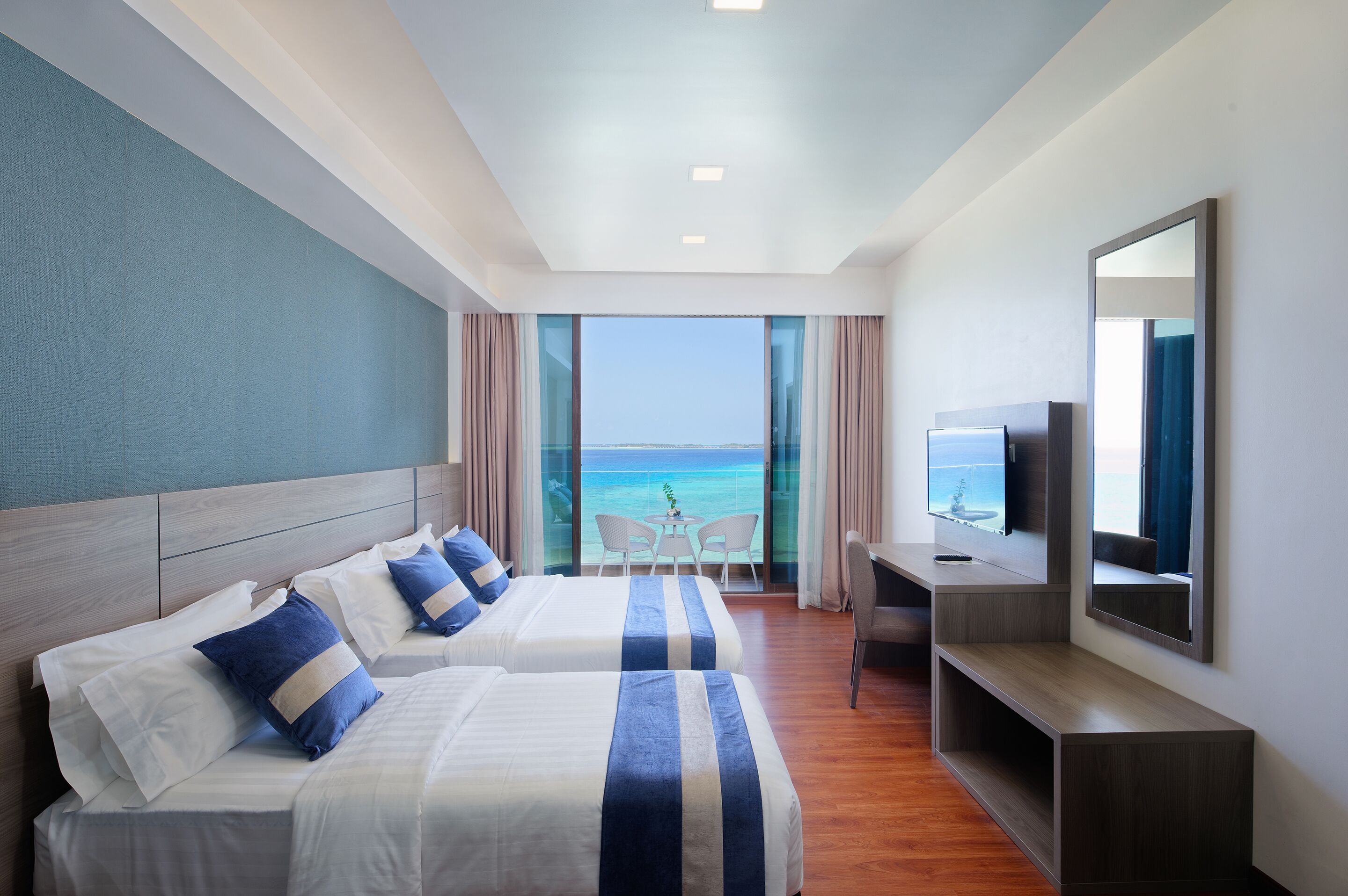 Habitación de hotel con dos camas, un escritorio, un televisor y un balcón con vista al mar.