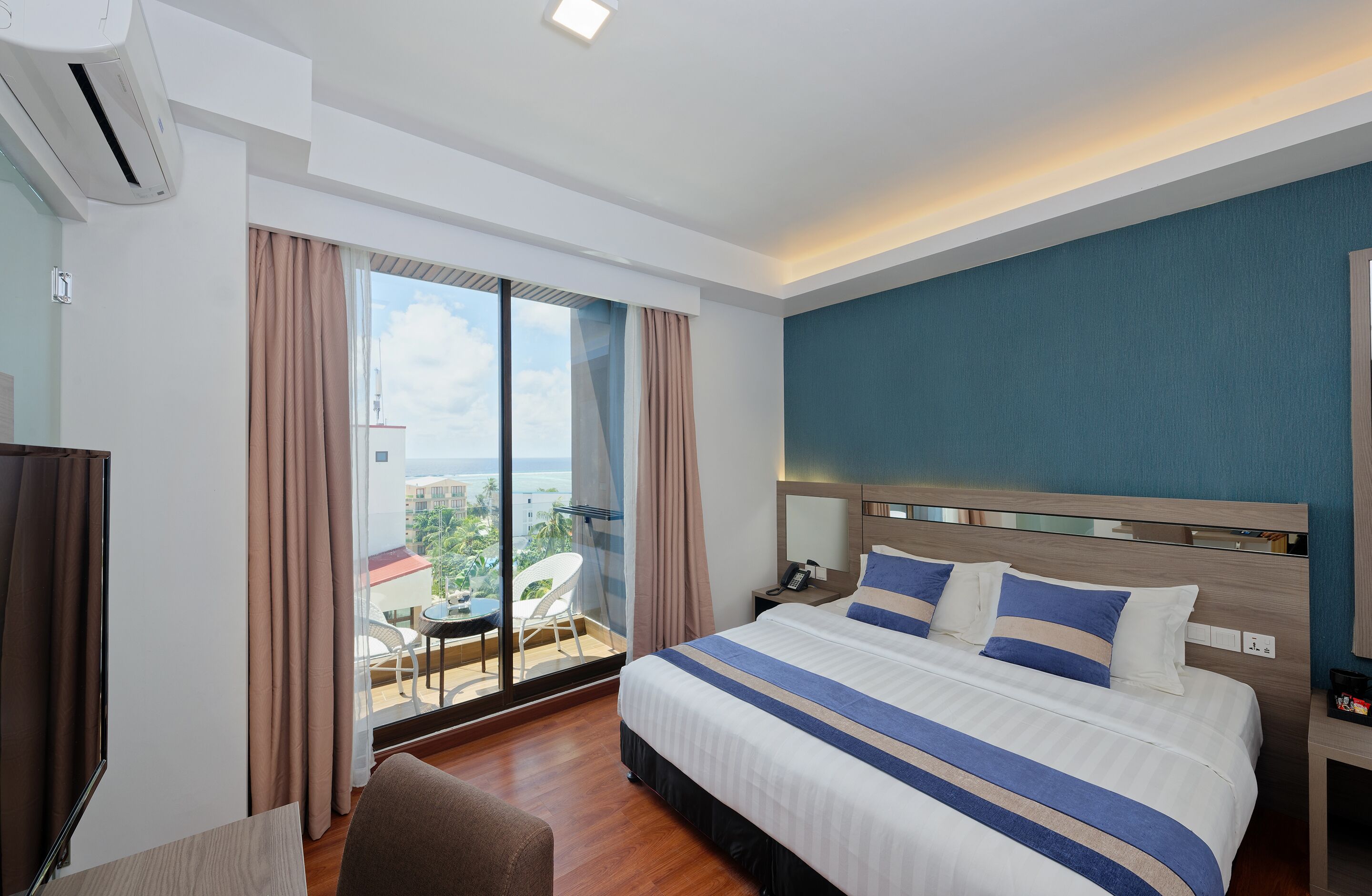 A hotel room with a large bed, a bedside table, a chair, and a view of the city through large windows.