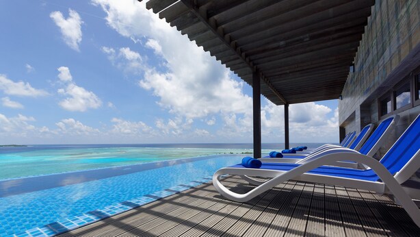 An infinity pool with clear blue water, lounge chairs, and a wooden deck. - Arena Beach Hotel (Maafushi)