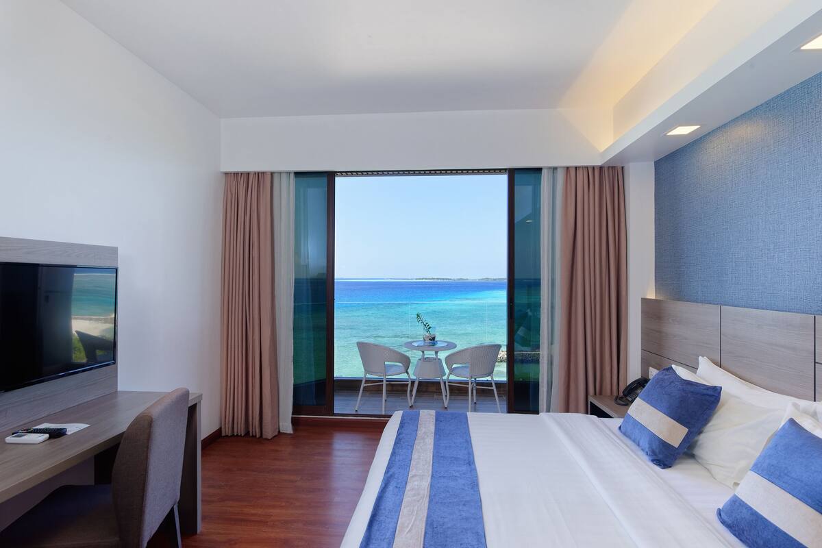 A hotel room with a large bed, a TV, a desk, and a balcony with a view of the ocean.