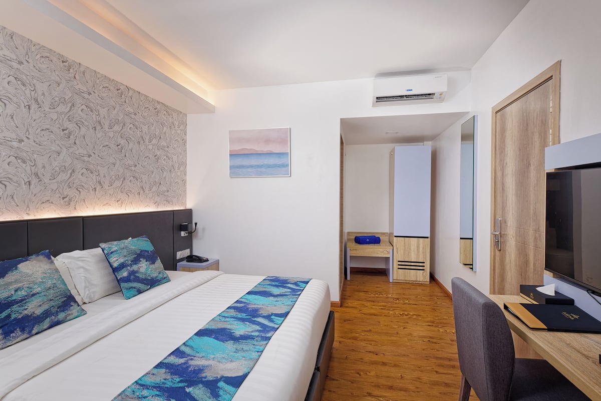 A modern hotel room with a large bed, a desk with a computer, a TV, and a bathroom visible in the background.