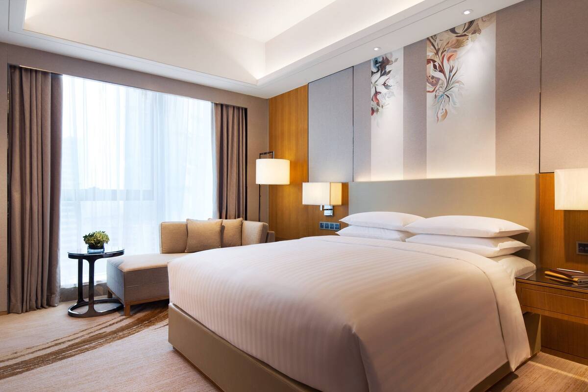 A hotel room with a large bed, a seating area with a chair and a small table, and a large window with curtains.