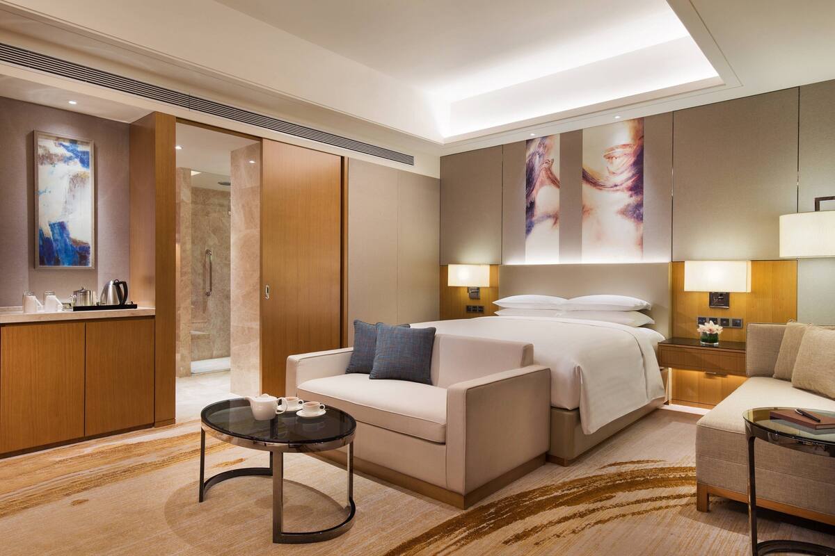 A modern hotel room with a bed, two sofas, a small table, and a desk with a lamp.