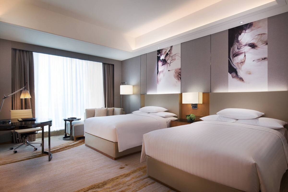 A hotel room with two beds, a desk, and a chair. There are large windows with curtains, a lamp, and artwork on the walls.