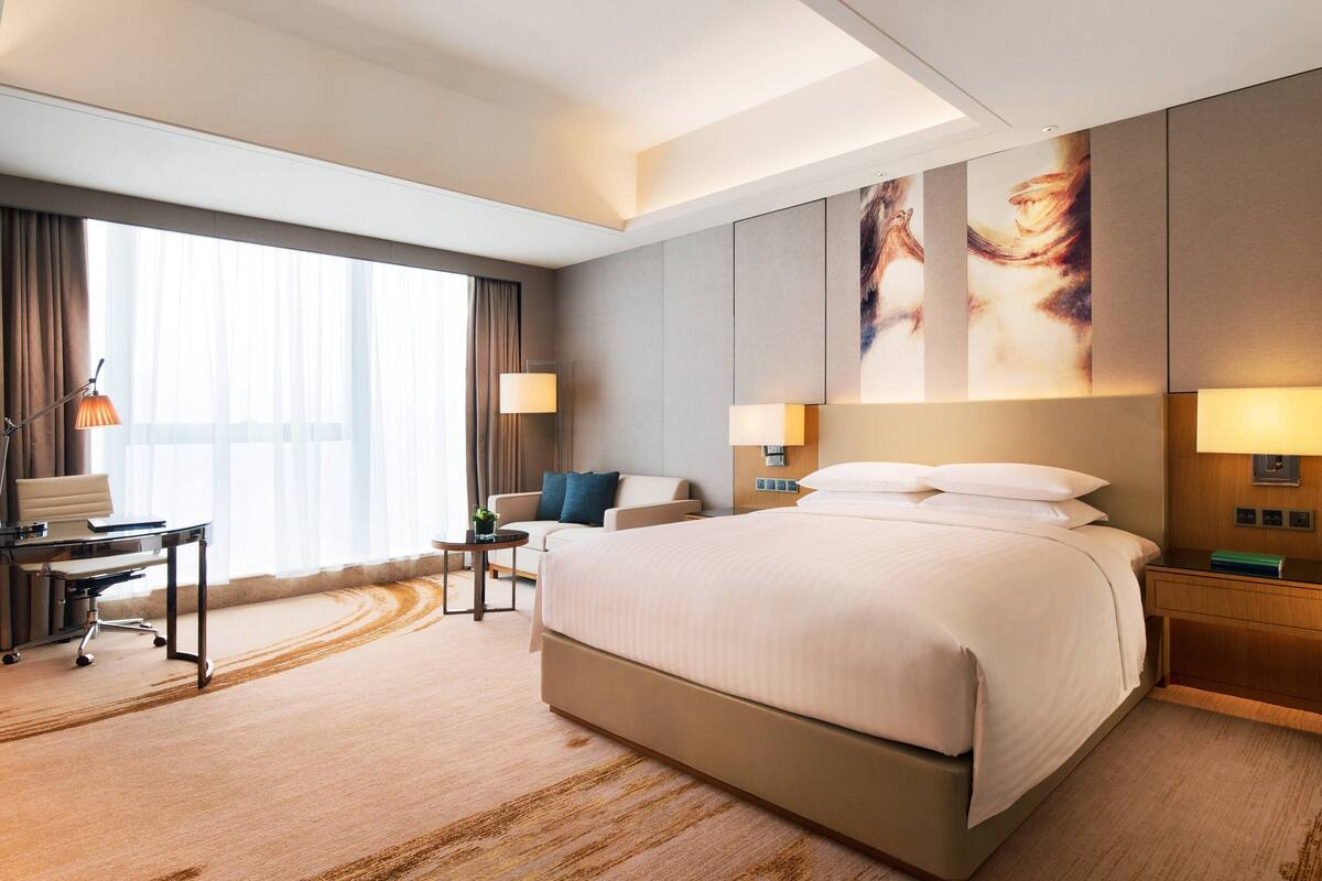 A hotel room with a large bed, a desk, a chair, and a sofa. There are two lamps on the nightstands and a window with curtains.