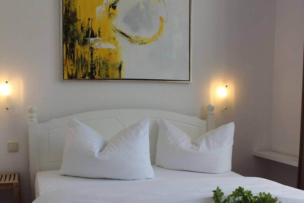 Un lit bien fait, avec du linge de lit blanc et des oreillers, une petite table de chevet avec une lampe, et un tableau accroché au-dessus du lit.