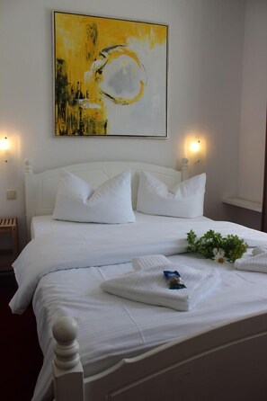 Ein ordentlich bezogenes Bett mit weißen Leinen und Kissen, ein kleiner Nachttisch mit Lampe sowie ein Gemälde über dem Bett.